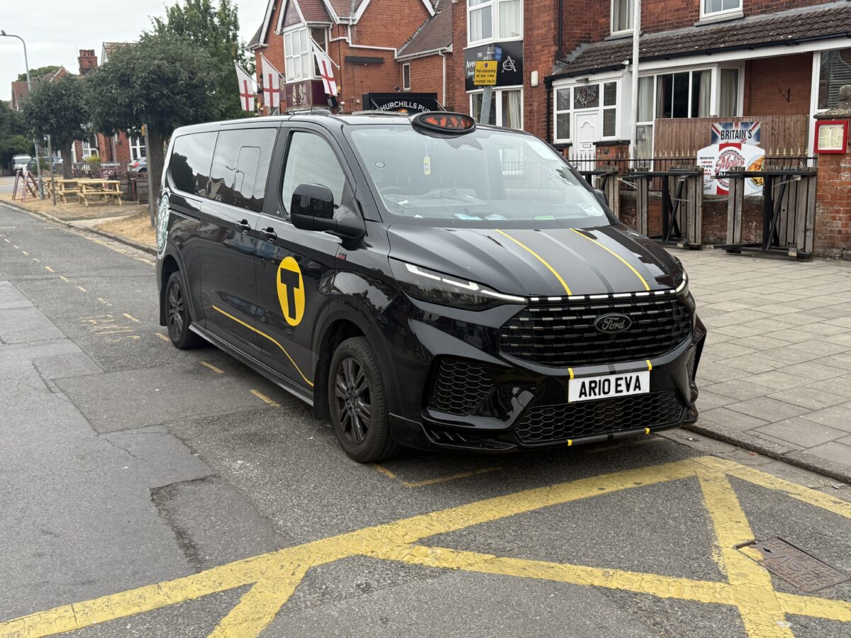 Black CJ’s Taxi long-wheelbase Ford Tourneo Custom 8-seater with yellow branding, viewed from the front and side.